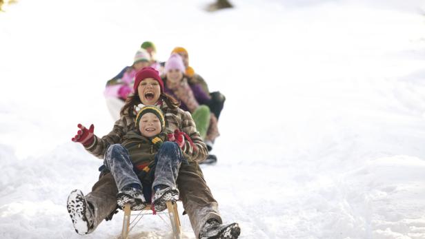 Eine Mutter und ihr Sohn rodeln mit einem Holzschlitten einen Hügel hinunter, gefolgt von anderen Rodlern.