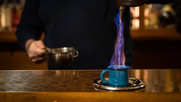 Ein Barkeeper gießt eine brennende Flüssigkeit in eine blaue Tasse.