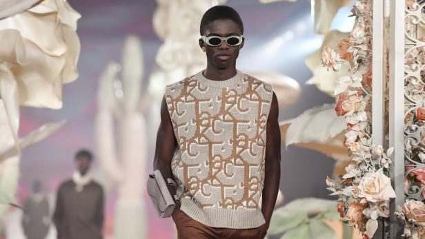 Ein dunkelhäutiges Model mit Sonnenbrille und Strickweste auf einem Laufsteg.