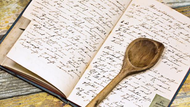 Ein altes Kochbuch liegt offen mit einem hölzernen Kochlöffel darauf.