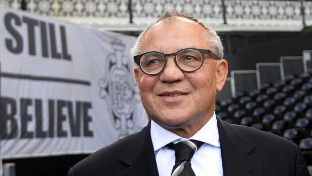 Ein Mann mit Brille und Anzug steht vor einer Tribüne mit einem Banner „Still Believe“.