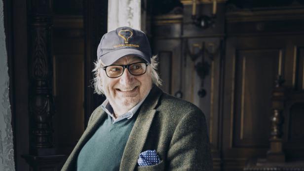 Ein lächelnder Mann mit Brille und Kappe posiert in einem eleganten Raum.