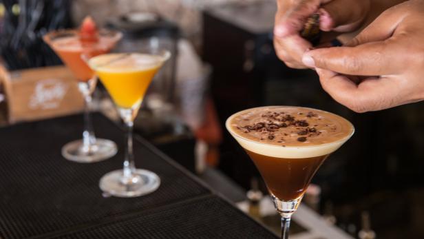 Ein Barkeeper garniert einen Espresso Martini mit Schokoladenraspeln. Im Hintergrund weitere Cocktails.