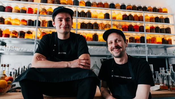 Zwei Männer in Kochkleidung posieren vor einem Regal voller Gläser mit Flüssigkeiten.