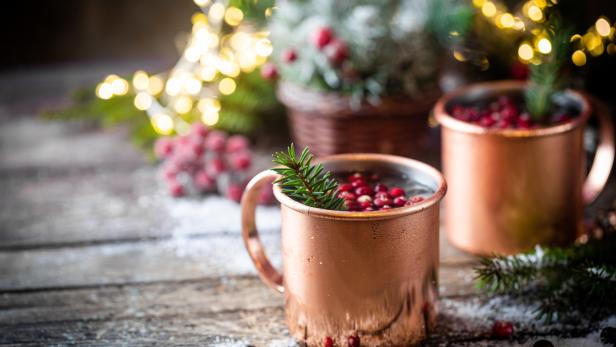 Zwei Kupferbecher mit Getränken und Preiselbeeren auf einem Holztisch mit weihnachtlicher Dekoration.