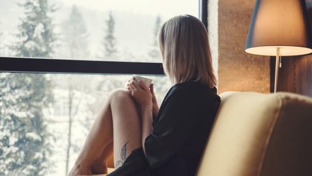 Eine Frau sitzt mit einer Tasse auf einem Sofa und blickt aus dem Fenster auf verschneite Bäume.