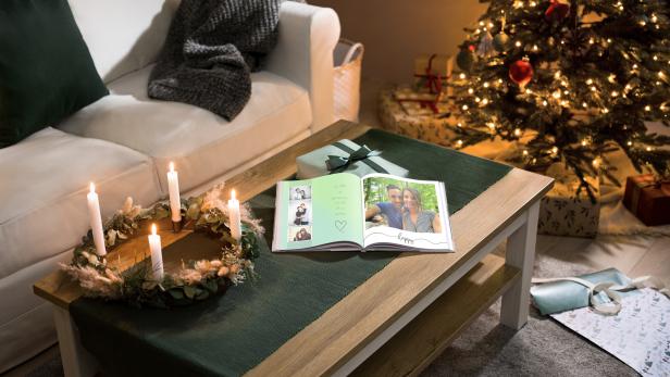 Ein Adventskranz mit brennenden Kerzen und ein geöffnetes Fotoalbum auf einem Tisch vor einem Weihnachtsbaum.