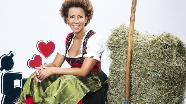 Eine Frau im Dirndl sitzt neben einem Heuballen und einer bemalten Kulisse mit Herzen.