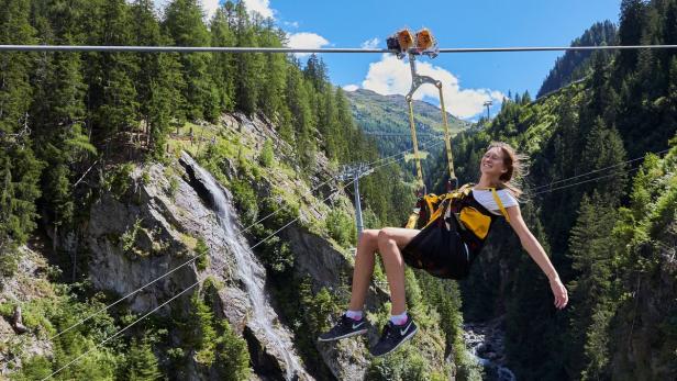 Eine junge Frau saust an einer Zipline durch eine grüne Berglandschaft mit Wasserfall.