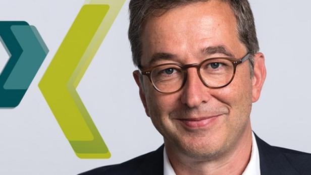 Ein lächelnder Mann mit Brille vor einem Logo mit grünen Pfeilen.