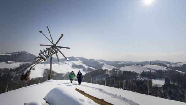 Zwei Frauen wandern im Winter durch eine verschneite Landschaft mit einem hölzernen Windrad.