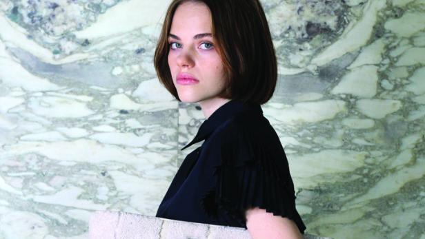 Eine Frau in einem schwarzen Kleid hält eine große, helle Tasche vor einem Marmorhintergrund.