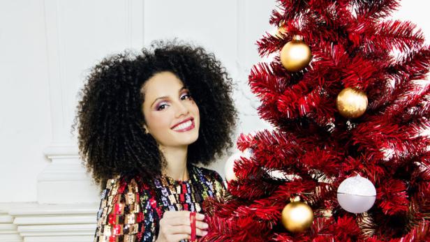 Eine Frau mit Afro-Haar und Paillettenkleid schmückt einen roten Weihnachtsbaum mit Kugeln.