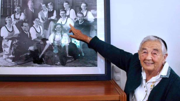 Eine ältere Frau zeigt auf ein Schwarzweißfoto einer Gruppe von Menschen in Tracht.