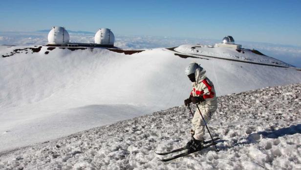 Ein Skifahrer fährt auf dem Mauna Kea in Hawaii Ski, mit Observatorien im Hintergrund.