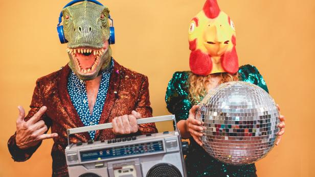 Eine Person mit Dinosaurier-Maske, Kopfhörern und Ghettoblaster neben einer Person mit Hühnermaske und Discokugel.