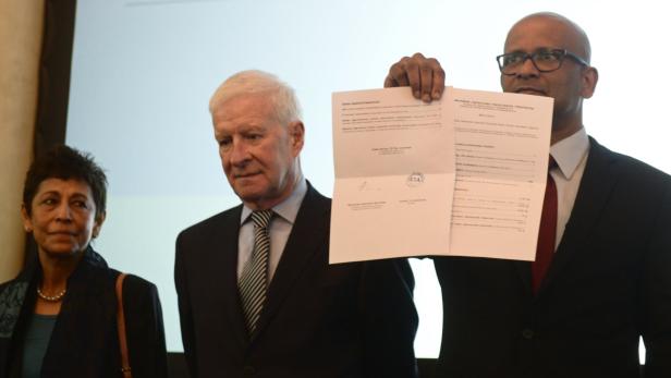 Drei Personen stehen, während ein Mann ein Dokument in die Kamera hält.