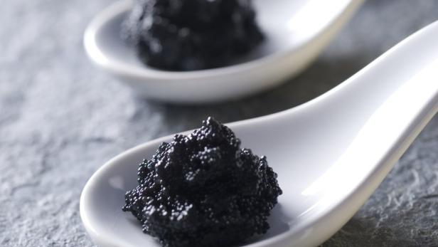 Drei weiße Löffel mit schwarzem Kaviar auf einer grauen Steinplatte.