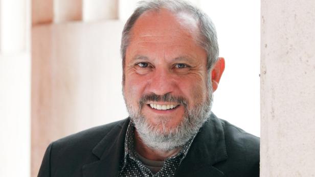 Ein lächelnder Mann mit grauem Bart und Sakko steht vor einer Wand.