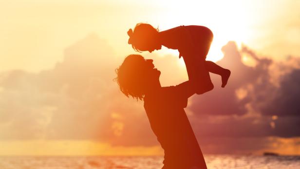 Ein Vater hält sein Kind am Strand in den Armen, im Hintergrund ein Sonnenuntergang.