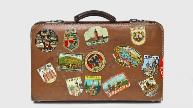 Ein brauner Koffer ist mit Aufklebern von verschiedenen Städten und Regionen beklebt, darunter Hamburg, Stockholm und Köln.