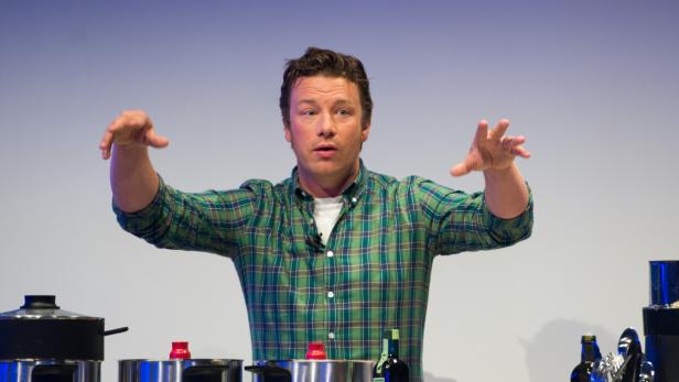 Jamie Oliver gestikuliert während einer Kochvorführung vor Töpfen.