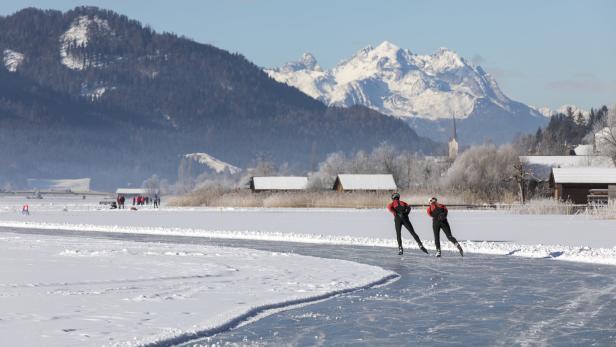 Zwei Personen laufen Schlittschuh auf einem zugefrorenen See vor einer Bergkulisse.