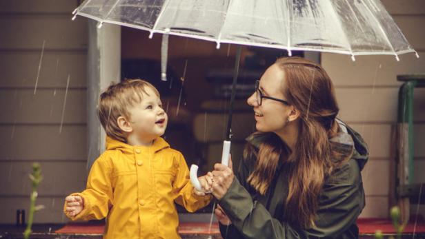 Eine Mutter und ihr Kind stehen mit einem Regenschirm im Regen vor einer Haustür.