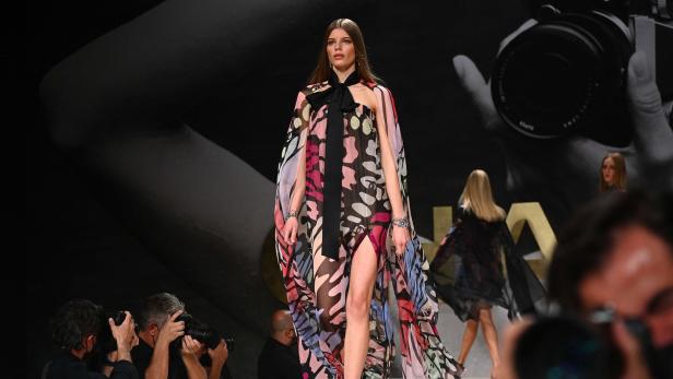 Ein Model präsentiert ein buntes, transparentes Kleid mit Schlitz auf einem Laufsteg.