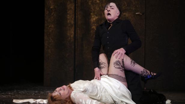 Auf einer Bühne liegt eine Frau im weißen Kleid, über ihr eine stehende Person in schwarzer Kleidung.