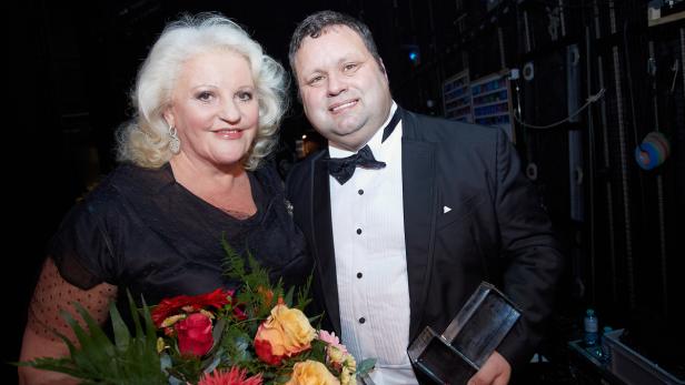 Eine Frau mit blonden Haaren und ein Mann im Smoking posieren mit einem Blumenstrauß und einer Trophäe.