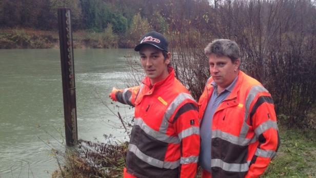 Zwei Männer in Warnkleidung stehen an einem Fluss und zeigen auf einen Pegelstandsanzeiger.