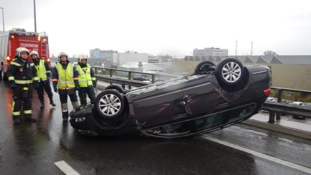 Ein auf dem Dach liegendes Auto auf einer Straße, mit Feuerwehrleuten im Hintergrund.