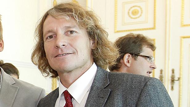 Ein Mann mit blonden, gewellten Haaren und einem grauen Sakko lächelt.