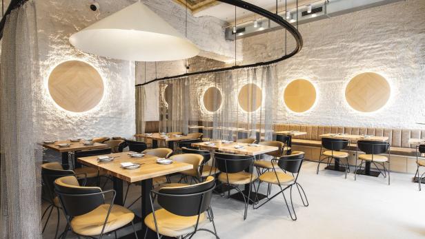 Innenansicht eines modernen Restaurants mit Holztischen und Stühlen vor einer strukturierten Wand mit kreisförmigen Leuchten.