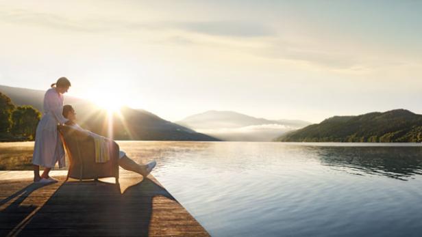 Eine Frau massiert einen Mann, der in einem Sessel auf einem Holzsteg an einem See sitzt.