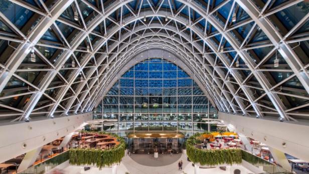 Die Lobby eines modernen Gebäudes mit Glasdach und begrünten Sitzbereichen.