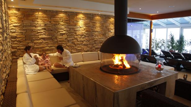 Eine Familie entspannt in Bademänteln vor einem Kamin in einem Wellnessbereich.