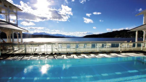 Ein Schwimmbad mit Blick auf einen See und bewaldete Hügel unter blauem Himmel.