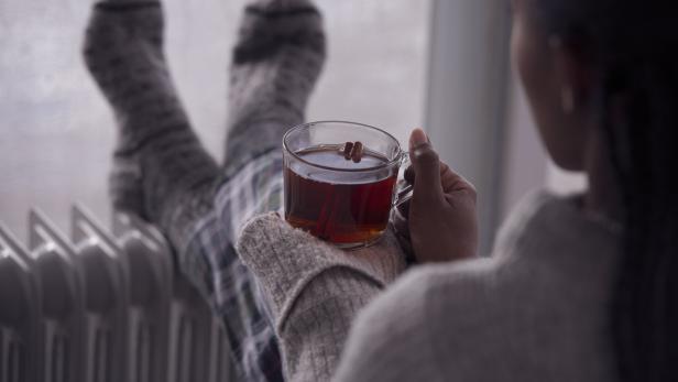 Eine Person mit Wollsocken wärmt sich mit einem Getränk vor einem Heizkörper.