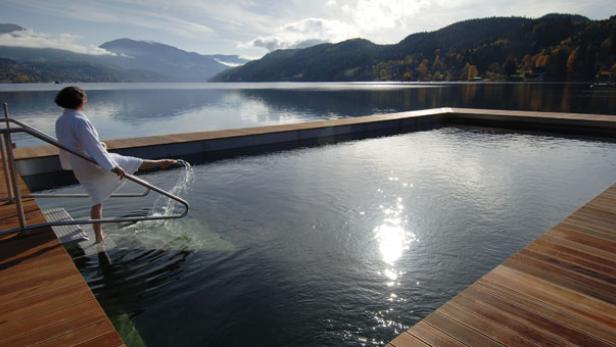 Eine Frau im Bademantel hält ihre Füße in einen Pool mit Seeblick.