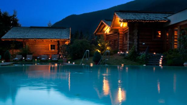 Ein beleuchteter Pool vor zwei Blockhäusern in einer Berglandschaft bei Dämmerung.