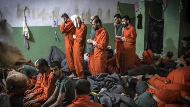 In einem überfüllten Raum sitzen und liegen Männer in orangefarbenen Overalls und grünen T-Shirts.