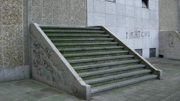 Eine graue Betontreppe vor einer Wand mit Graffiti, darunter der Schriftzug „Pikatchu“.