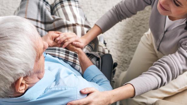 Eine junge Frau hält die Hand eines älteren Mannes im Rollstuhl.