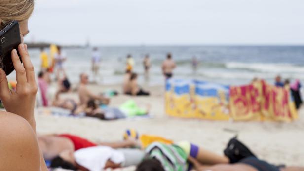 Eine Frau telefoniert am Strand, während andere sich sonnen und im Meer baden.