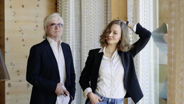 Ein Mann mit Sonnenbrille und Zigarre posiert mit einer jungen Frau vor einem Fenster.