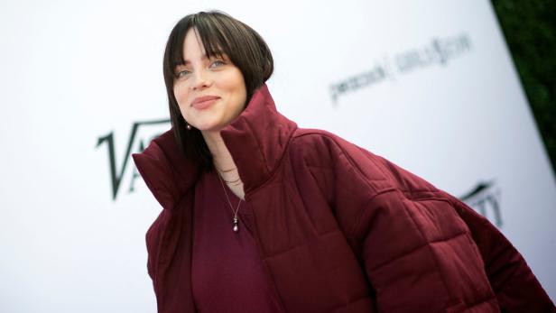 Billie Eilish trägt eine weinrote Jacke und steht vor einem weißen Hintergrund.