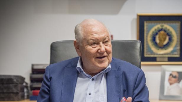 Ein älterer Mann mit blauem Sakko sitzt an einem Schreibtisch.