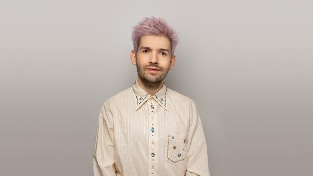 Ein Mann mit rosa Haaren und einem verzierten Hemd blickt in die Kamera.